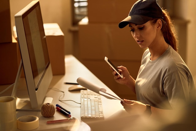 Joven repartidora usando teléfono móvil mientras revisa la lista de despacho en la oficina