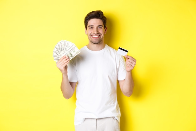 Joven retirar dinero de la tarjeta de crédito, sonriendo complacido, de pie sobre fondo amarillo.