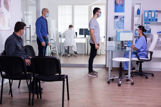 Joven revisando la cita respetando el distanciamiento social en la sala de espera del hospital, la enfermera busca en la computadora con máscara facial contra el covid-19