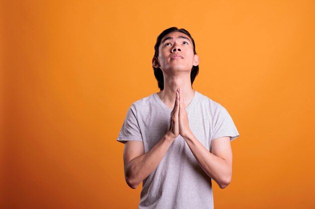 Joven rezando manos plegables, fe, concepto de adoración. Adolescente asiático religioso suplicando, mirando hacia arriba con esperanza, persona alabando a los dioses, plano medio sobre fondo naranja
