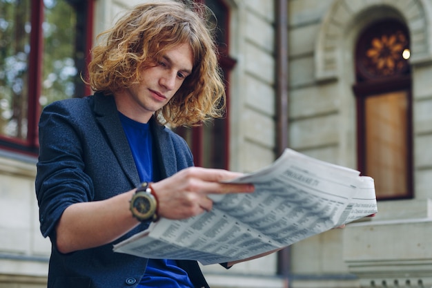 Joven rojizo leyendo periódico cerca de edificio de estilo antiguo