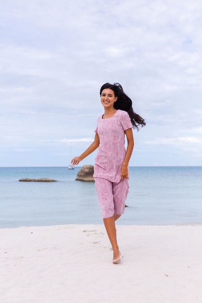 Joven romántica con cabello largo y oscuro en un vestido en la playa sonriendo y riendo pasando un rato agradable solo