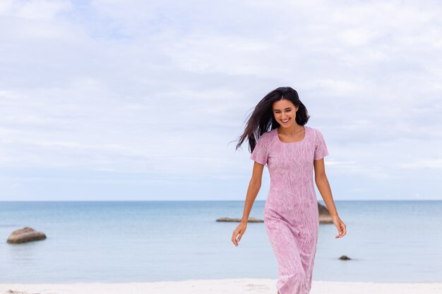 Joven romántica con cabello largo y oscuro en un vestido en la playa sonriendo y riendo pasando un rato agradable solo