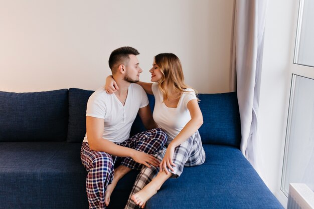 Joven romántico disfrutando de fin de semana con su esposa. Hermosa chica con cabello rubio escalofriante en el sofá con su novio.