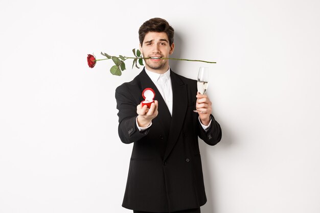 Joven romántico en traje haciendo una propuesta, sosteniendo una rosa en los dientes y una copa de champán, mostrando el anillo de compromiso, pidiendo casarse con él, de pie contra el fondo blanco.