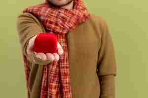 Foto gratuita joven en ropa casual con bufanda alrededor del cuello mostrando caja roja con anillo de compromiso concepto del día de san valentín de pie sobre la pared verde