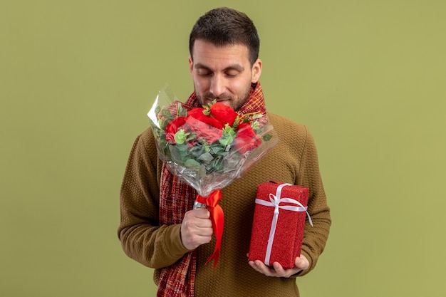 Joven en ropa casual con bufanda alrededor del cuello sosteniendo un ramo de rosas rojas y presente feliz y positivo concepto de día de San Valentín sonriente de pie sobre fondo verde