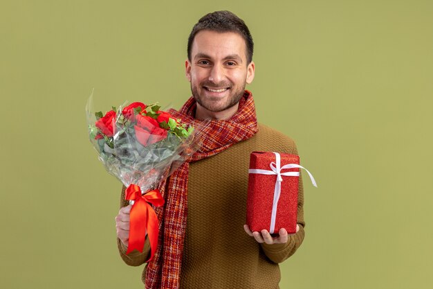 Joven en ropa casual con bufanda alrededor del cuello sosteniendo un ramo de rosas rojas y presente mirando a la cámara feliz y alegre concepto de día de San Valentín sonriente de pie sobre fondo verde