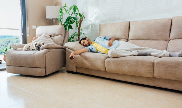 Joven en ropa casual durmiendo en un sofá en casa con un control remoto de TV en la mano