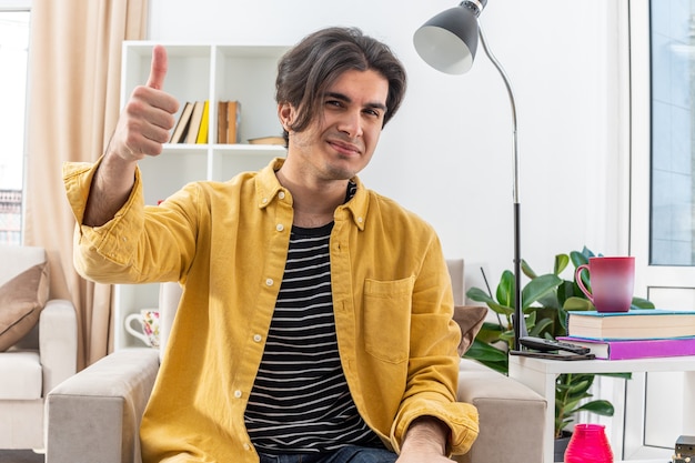 Joven en ropa casual sonriendo alegremente mostrando los pulgares para arriba sentado en la silla en la sala de luz