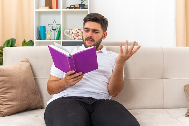 Joven en ropa casual sosteniendo un libro leyendo con expresión confusa sentado en un sofá en la sala de estar luminosa