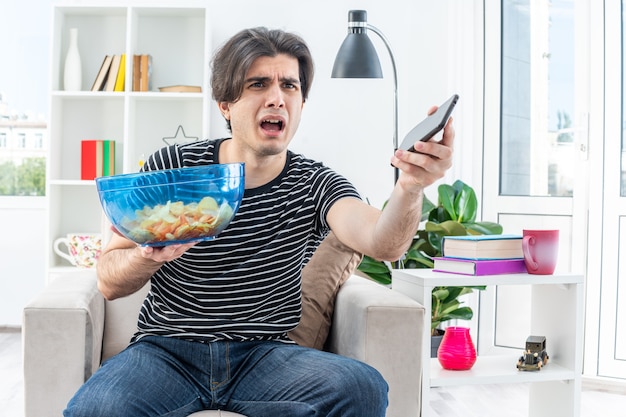 Joven en ropa casual sosteniendo un tazón de papas fritas y un teléfono inteligente mirando a un lado confundido y disgustado sentado en la silla en la sala de estar luminosa