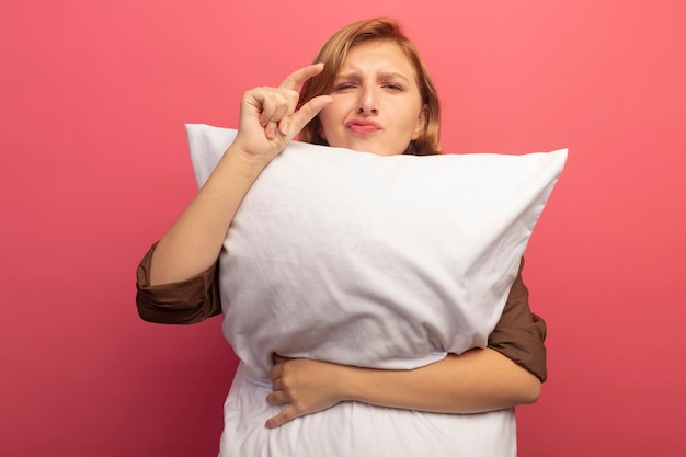 Foto gratuita joven rubia abrazando la almohada mirando haciendo gesto de pequeña cantidad aislado en la pared rosa