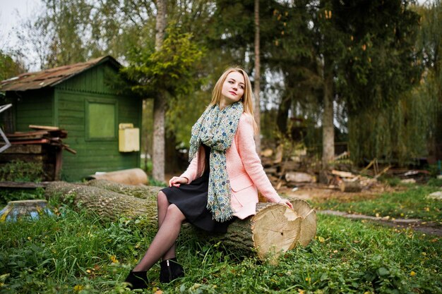 Joven rubia con abrigo rosa posada en el parque de otoño sentada en un árbol cortado contra la casa del guardabosques