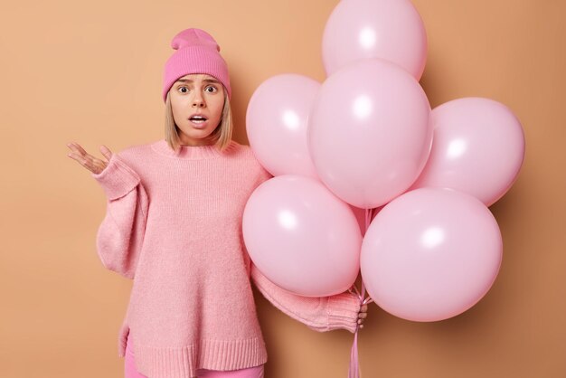 Una joven rubia avergonzada usa un puente casual y poses de sombrero con globos de helio sorprendida al escuchar malas noticias celebra el aniversario aislado sobre fondo marrón. Concepto de celebración de cumpleaños