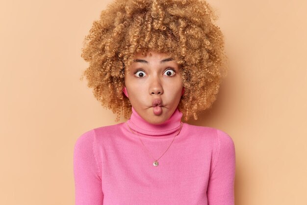 Una joven rubia de cabello rizado, graciosa y tonta, hace que los labios de pescado se plieguen, mira fijamente, sorprendida, hace que la expresión infantil use un cuello de tortuga rosa aislado sobre un fondo marrón. Chica hace ejercicios faciales