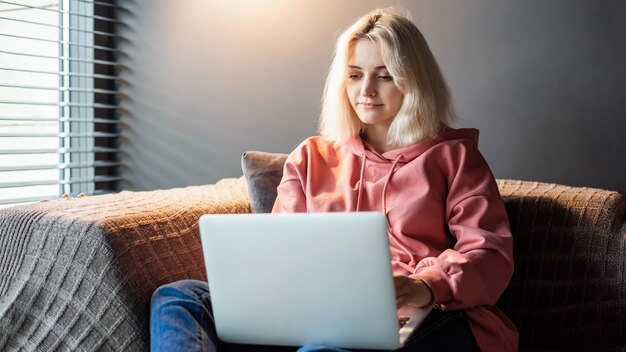 La joven rubia creadora de contenido está en su computadora portátil sentada en el sofá cerca de la ventana. Trabajando desde casa