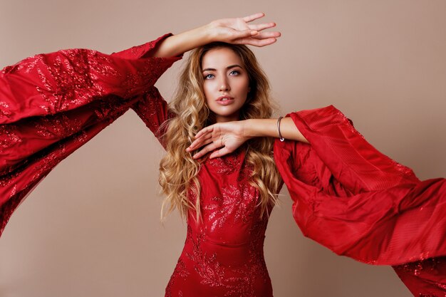 joven rubia encantadora en lujoso vestido rojo con mangas anchas. Pose expresiva.