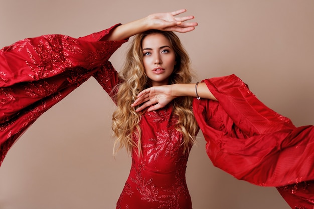 joven rubia encantadora en lujoso vestido rojo con mangas anchas. Pose expresiva.