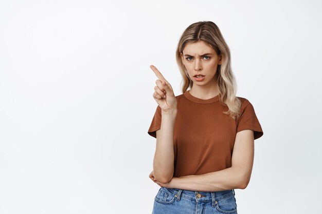 Joven rubia enojada señalando con el dedo la esquina superior izquierda de las cejas y luciendo disgustada de pie en camiseta contra fondo blanco