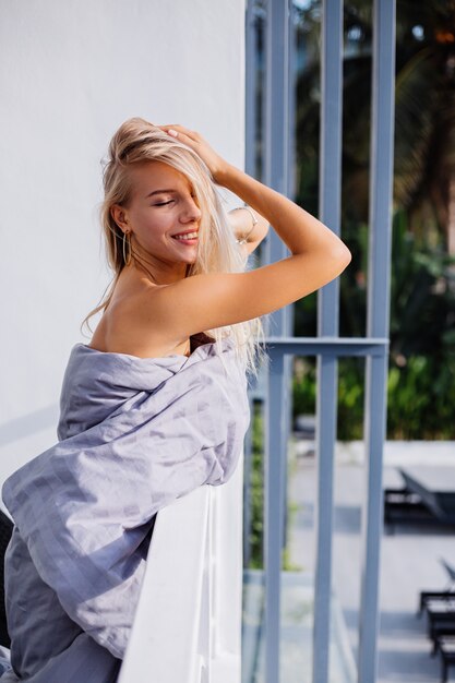 Joven rubia con estilo europeo en manta en el balcón tropical se encuentra con el amanecer en la mañana.