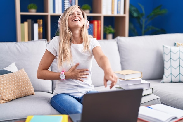 Joven rubia estudiando usando una computadora portátil en casa riéndose de ti, señalando con el dedo a la cámara con la mano sobre el cuerpo, expresión de vergüenza