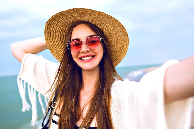Joven rubia feliz haciendo selfie, con sombrero de paja y gafas de sol lindas de corazón, disfruta de sus vacaciones de verano cerca del océano.