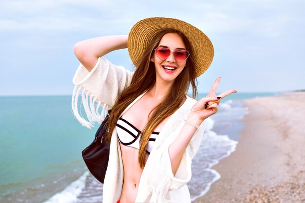 Joven rubia feliz posando en la playa, con sombrero de paja y gafas de sol lindas de corazón, disfruta de sus vacaciones de verano cerca del océano, vistiendo bikini y chaqueta boho.