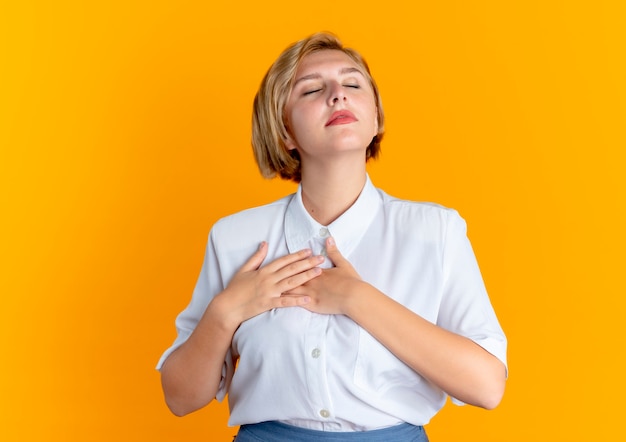 Joven rubia feliz rusa pone las manos en el pecho con los ojos cerrados aislado sobre fondo naranja con espacio de copia