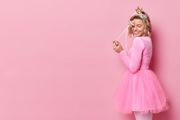 Una joven rubia feliz viste un vestido festivo y finge ser un hada mágica que sostiene una varita de perfil aislada sobre un espacio vacío de fondo rosa para tu contenido promocional Princesa en la fiesta
