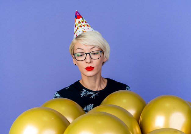 Joven rubia fiestera con gafas y gorro de cumpleaños de pie detrás de globos mirándolos aislado sobre fondo púrpura