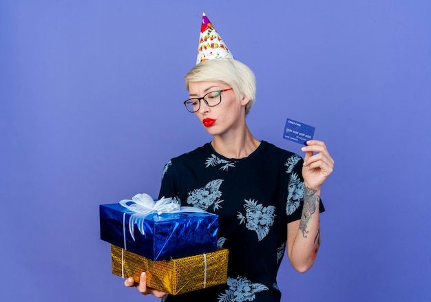 Joven rubia fiestera con gafas y gorro de cumpleaños sosteniendo cajas de regalo y tarjeta de crédito mirando cajas aisladas sobre fondo púrpura con espacio de copia