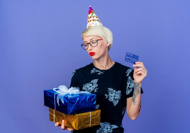 Joven rubia fiestera con gafas y gorro de cumpleaños sosteniendo cajas de regalo y tarjeta de crédito mirando cajas aisladas sobre fondo púrpura con espacio de copia