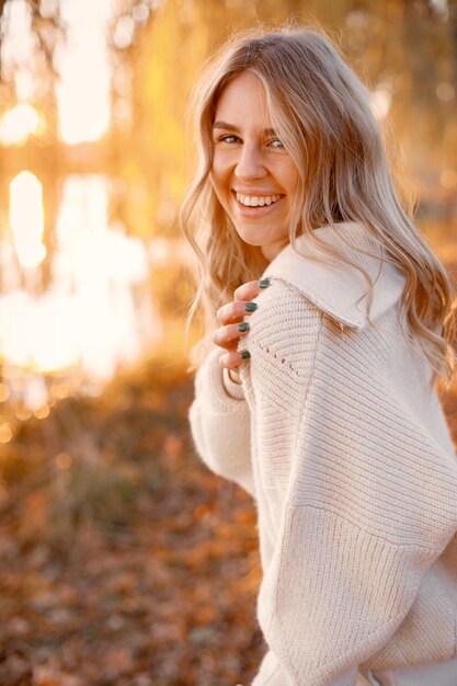 Joven rubia con flores de pie en el parque de otoño cerca del lago Mujer con suéter beige Chica posando para una foto en un día soleado