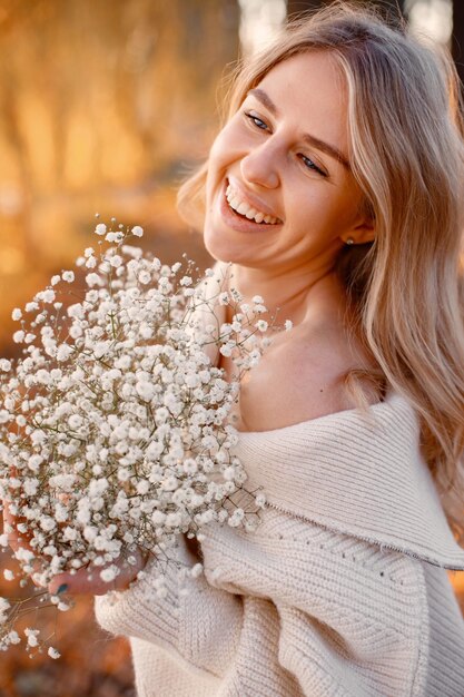 Joven rubia con flores de pie en el parque de otoño cerca del lago Mujer con suéter beige Chica posando para una foto en un día soleado
