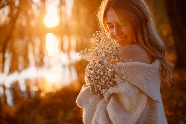 Joven rubia con flores de pie en el parque de otoño cerca del lago Mujer con suéter beige Chica posando para una foto en un día soleado