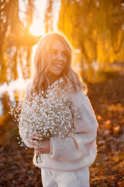 Joven rubia con flores de pie en el parque de otoño cerca del lago Mujer con suéter beige y pantalones blancos Chica posando para una foto en un día soleado
