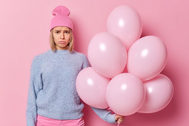 Una joven rubia frustrada e infeliz mira con tristeza a la cámara que sostiene un gran montón de globos inflados y se siente sola durante la celebración del cumpleaños vestida con ropa informal aislada en un fondo rosado