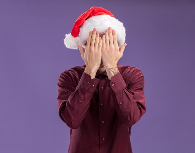 Joven rubia con gorro de Papá Noel y gafas que cubren la cara con las manos aisladas sobre fondo púrpura con espacio de copia