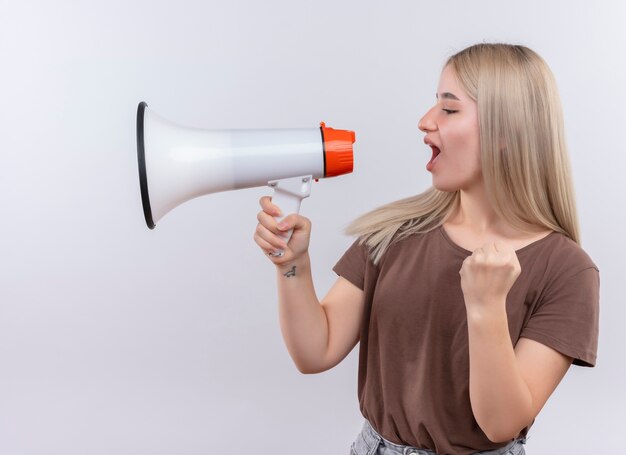 Joven rubia hablando por el altavoz con el puño levantado en la pared blanca aislada