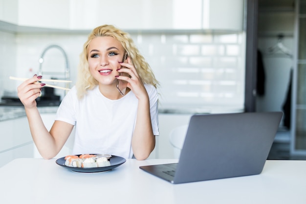 Joven rubia hablando por teléfono sentado en su cocina comer sushi en casa mesa trabajando con una computadora portátil en la mañana