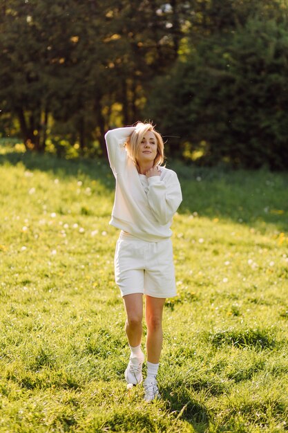 Joven rubia lleva una sudadera con capucha blanca sonriendo y caminando en el bosque