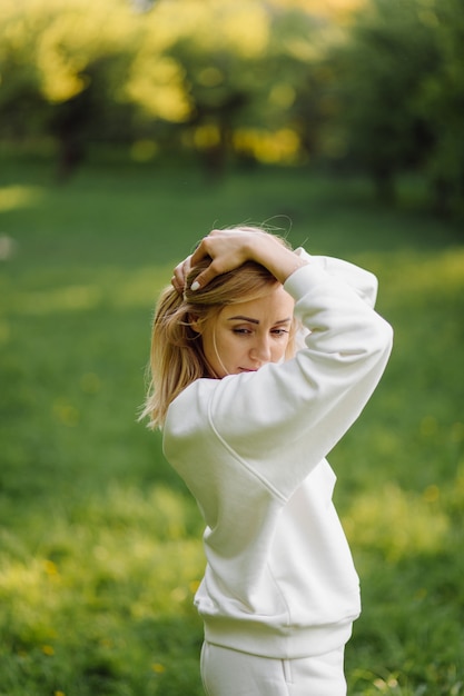 Joven rubia lleva una sudadera con capucha blanca sonriendo y caminando en el bosque