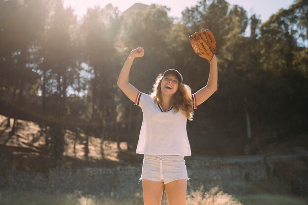 Joven rubia con las manos levantadas y el guante de béisbol en el fondo de la naturaleza