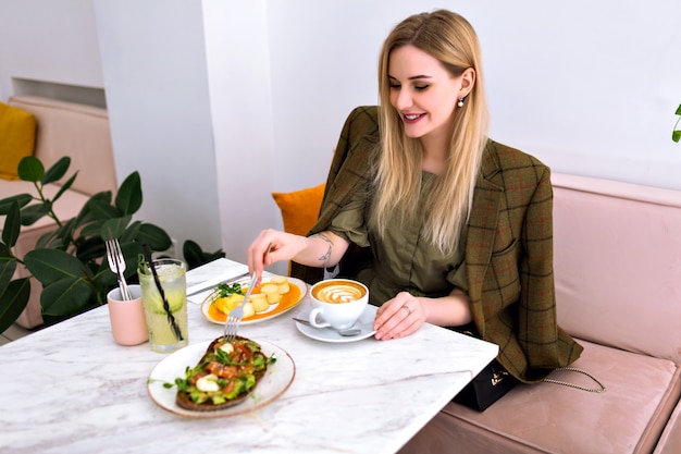 Joven rubia muy sonriente disfrutando de un sabroso brunch saludable con tostadas de salmón y aguacate, capuchino, limonada y postre, traje elegante, interior elegante y ligero.