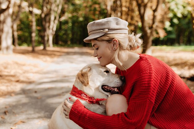 Joven rubia en ropa roja de moda sonriendo a su hermoso labrador en el parque de otoño. Hermosa chica y su perro se relajan al aire libre.