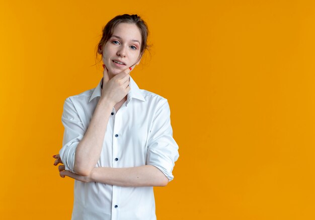 Joven rubia rusa confiada pone la mano en la barbilla aislada en el espacio naranja con espacio de copia