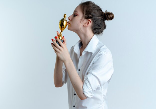 Joven rubia rusa confiada sostiene y besa la copa ganadora aislada en el espacio en blanco con espacio de copia