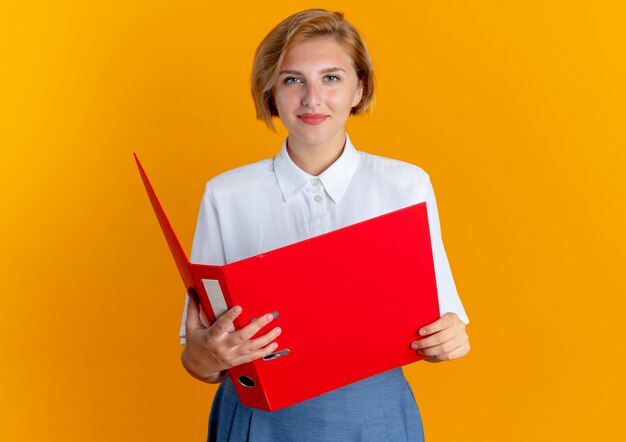 Joven rubia rusa contenta tiene carpeta de archivo mirando a cámara aislada sobre fondo naranja con espacio de copia