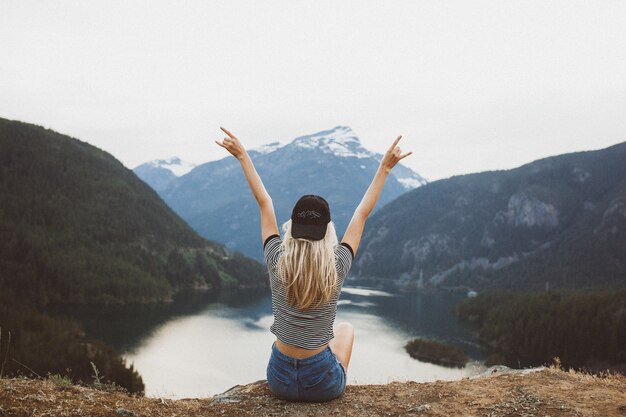 Joven rubia sentada en el acantilado disfrutando de la vista de las montañas y el lago